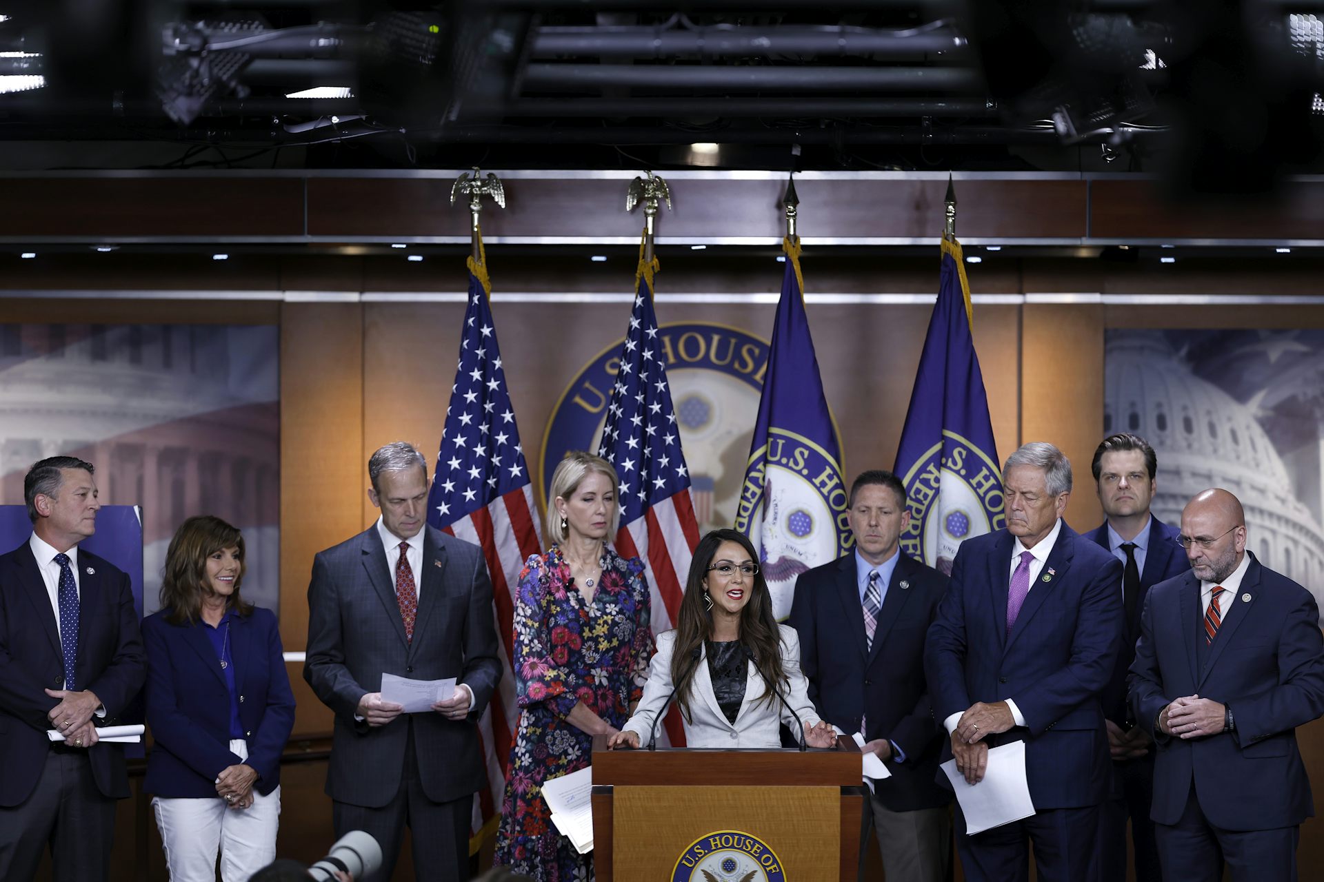 A woman with long dark hair, wearing eye glasses and a white suit speaks from a lectern. Standing behind her are suited men and women, some holding papers in their hands.