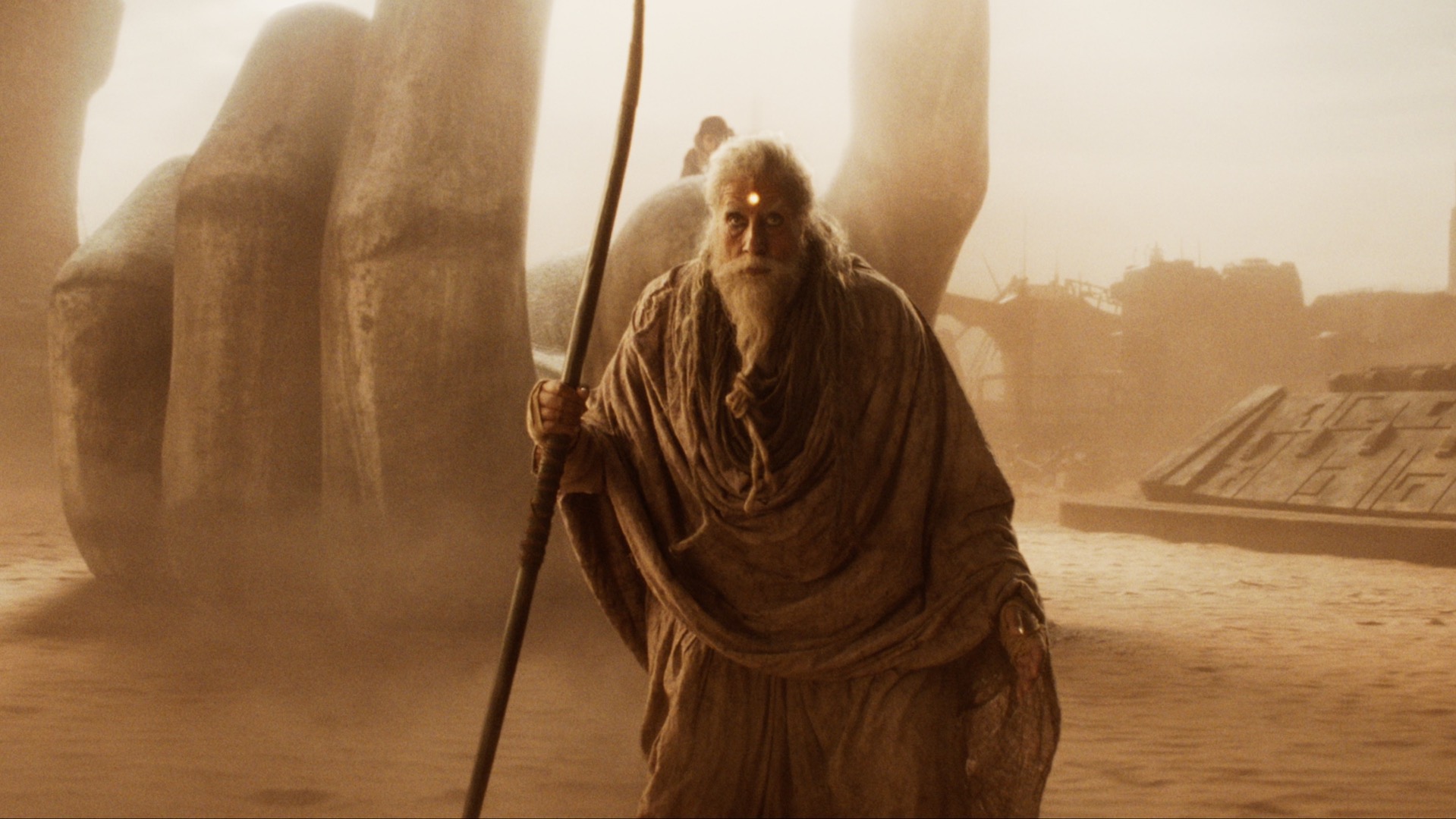 An old man dramed in robes pictured in a desert scape and holding a staff; Amitabh Bachchan in 'Kalki 2898 AD'