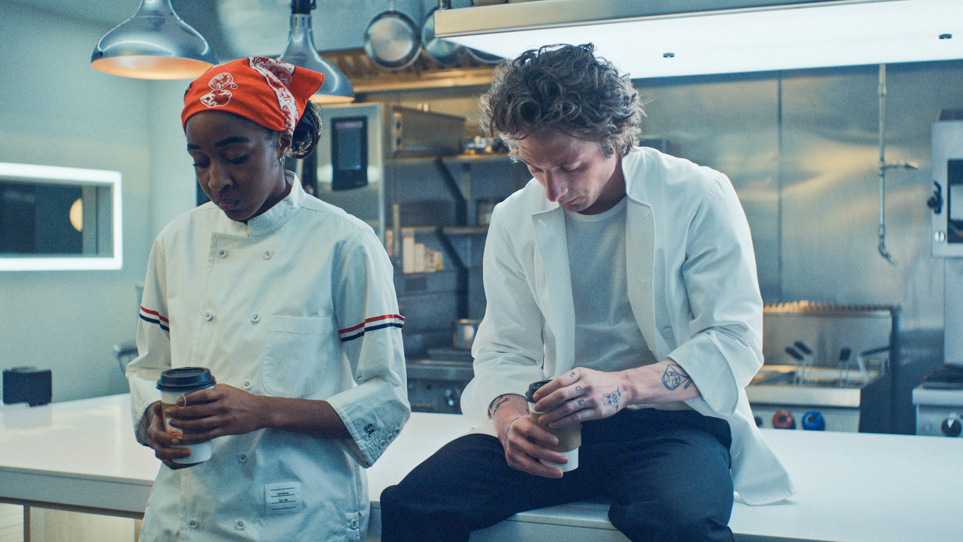 Two chefs with coffee at a kitchen island, one sitting atop and the other leaning back against it; still from 'The Bear'