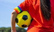 Individual in a orange sports top holding a football under their arm with a rainbow armband