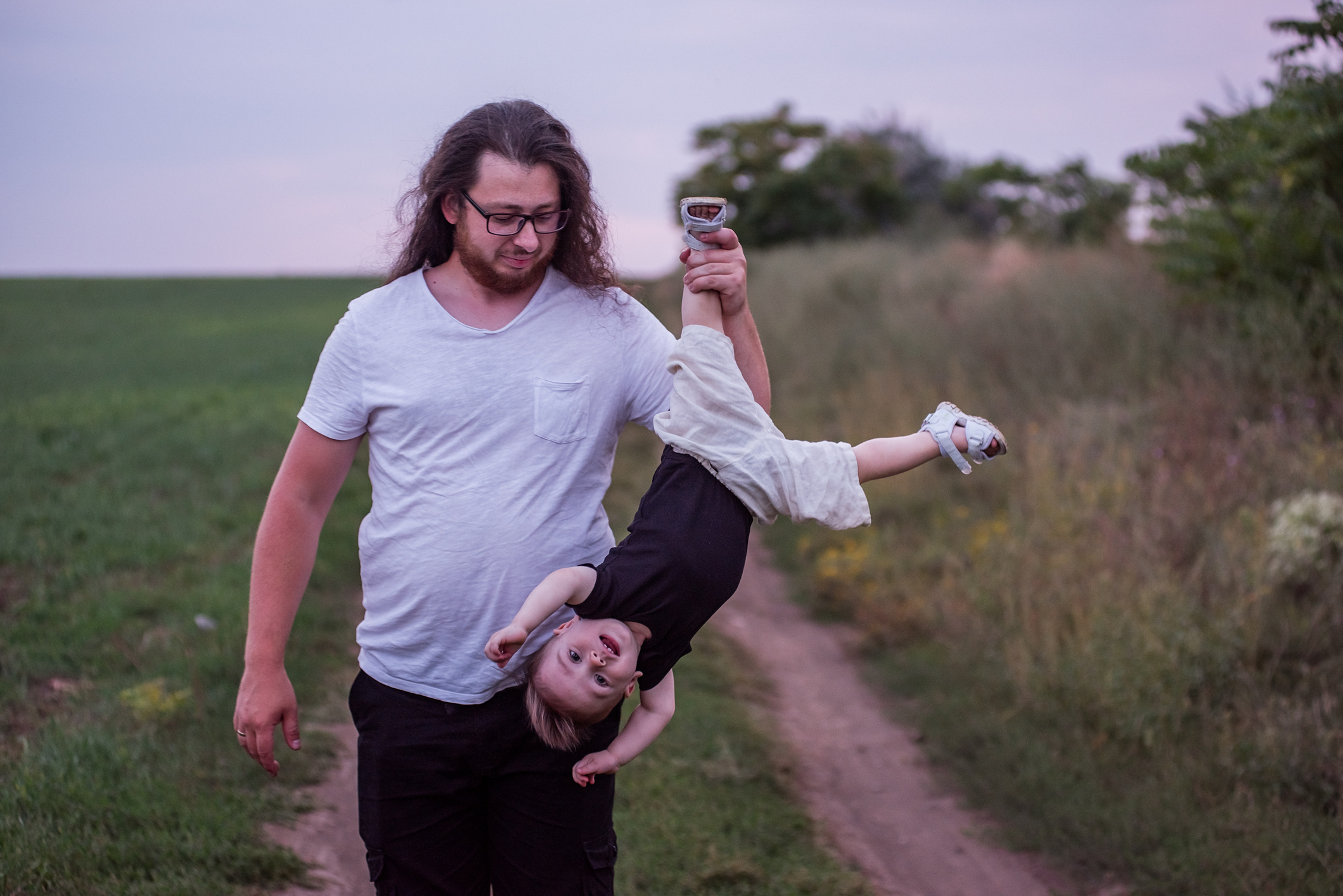 Diverse father with long curly hair, beard, and glasses holding his toddler son playfully upside down in a green field at sunset