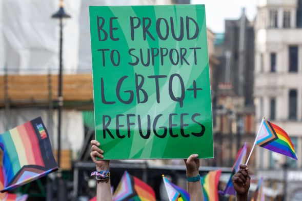 This is an image of a sign in black text on a green board. The text reads "Be Proud to support LGBTQ+ refugees". THere are various pride flags in hand