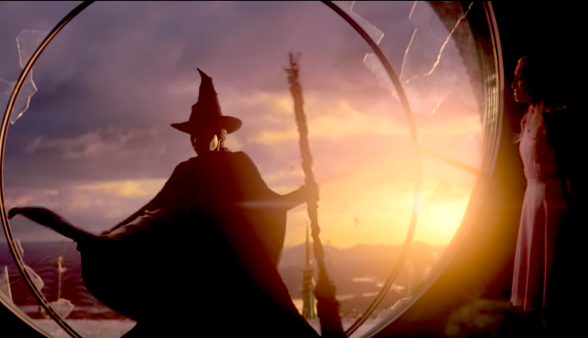 Screenshot from Wicked trailer showing Elphaba in her trademark pointed witch hat and cape and broomstick, about to leap from a broken window. The sun is setting behind her