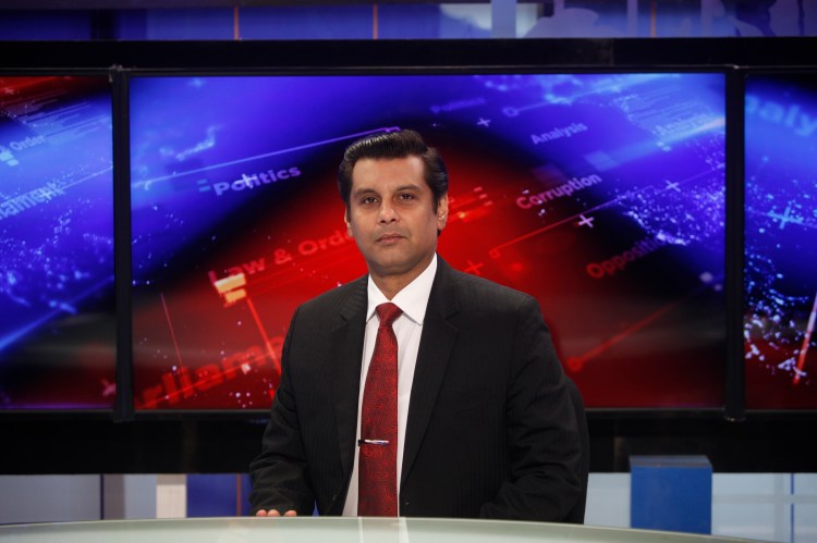A man in a suit stands before a news broadcast background.