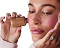 A woman with freckles examines a bottle of foundation, holding it close to her face with manicured h...