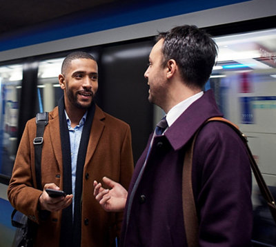 Dois homens conversando em uma plataforma de metrô, com um segurando um smartphone e um trem desfocado em movimento atrás deles.