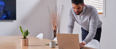 Um homem de suéter cinza em pé e inclinado sobre um laptop em uma mesa de madeira em um escritório iluminado.