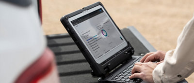Uma pessoa trabalha em um laptop robusto exibindo gráficos, sentado ao ar livre em uma superfície de surface arenosa.