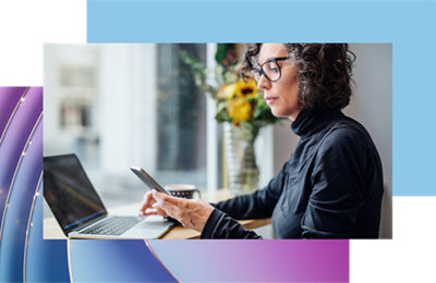 Mulher de óculos usando um smartphone e um laptop em uma mesa de café com flores e xícara de café por perto, parecendo concentrada.