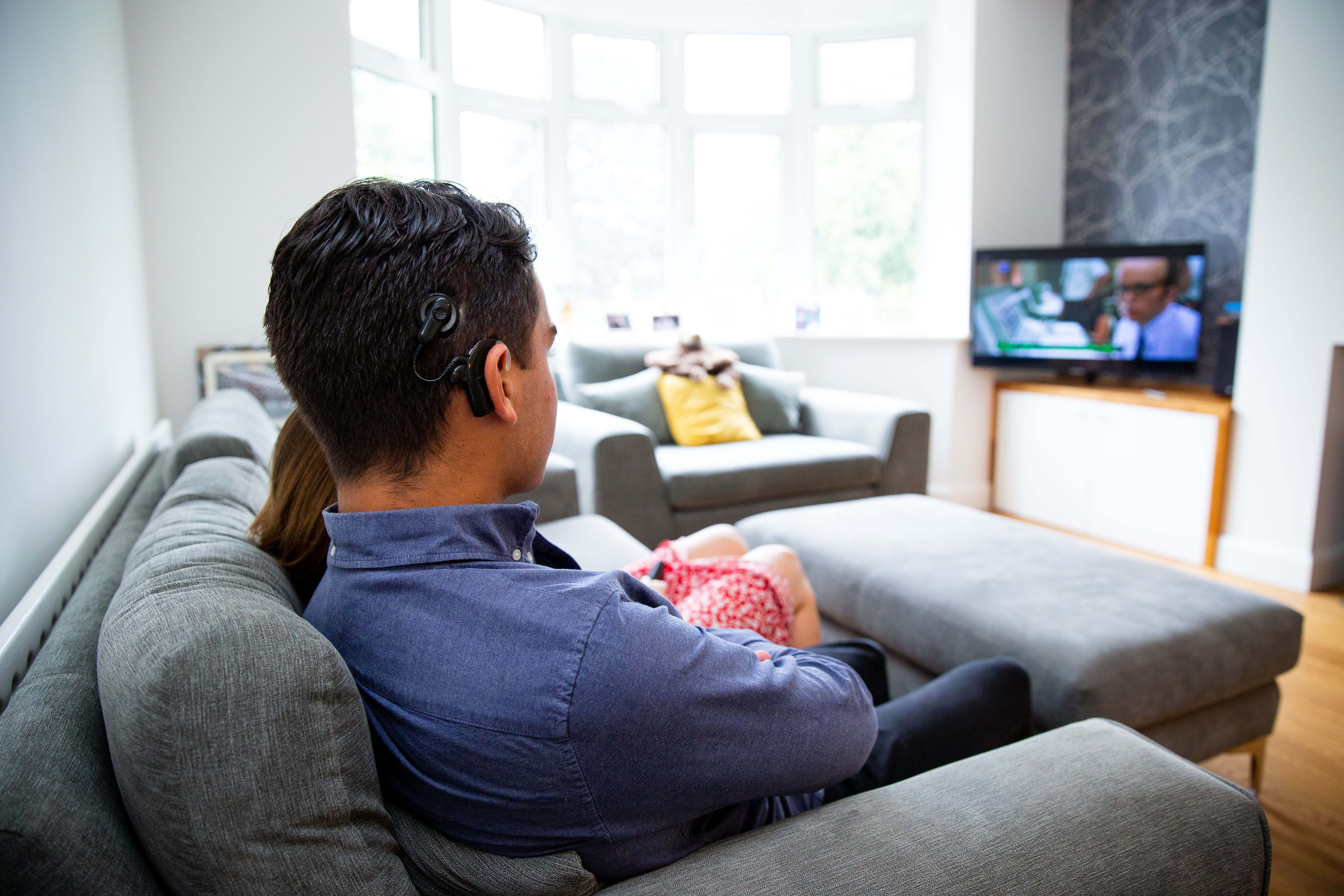 Photo taken over the shoulder over a young man wearing a blue shirt and a cochlear implant. He sits on the sofa beside a woman wearing a red skirt, watching TV with subtitles on.