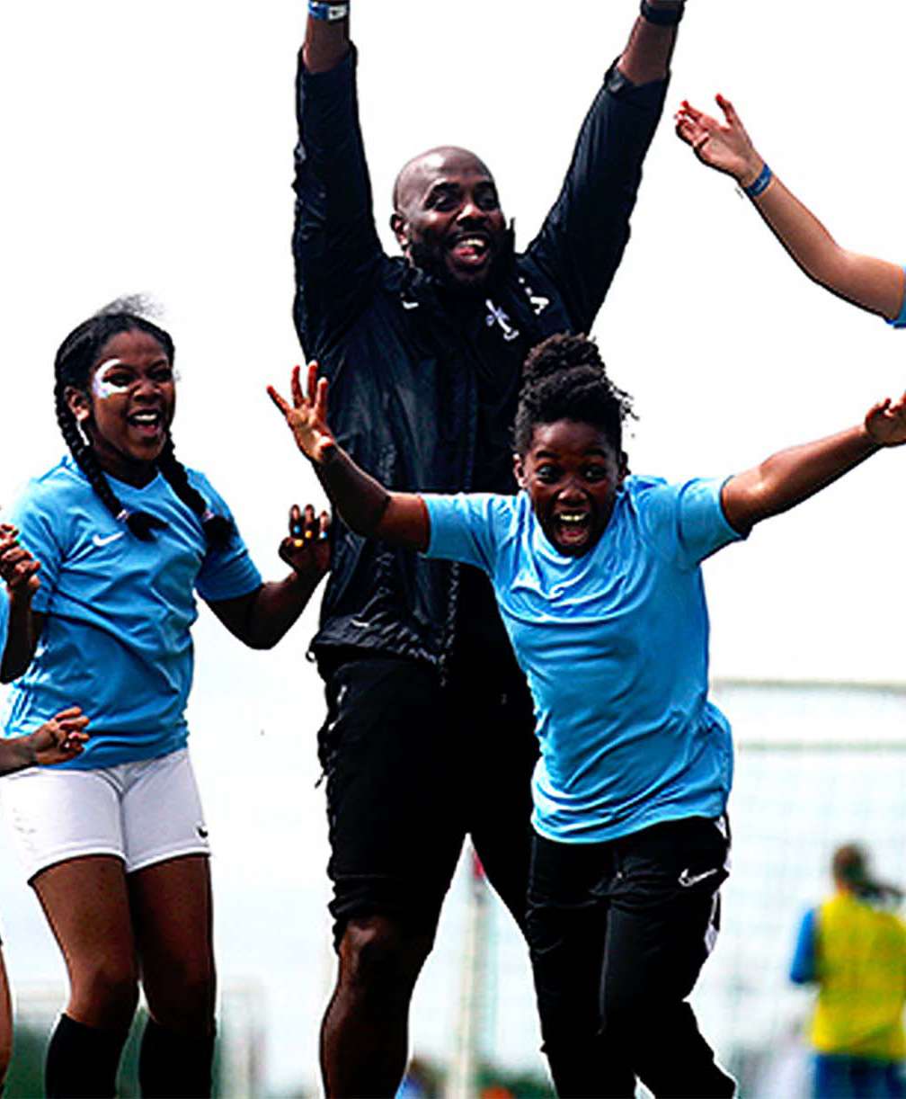7 children stand in front of a man, all celebrating with their hands in the air. The children wear blue football tops and the man wears a black tracksuit