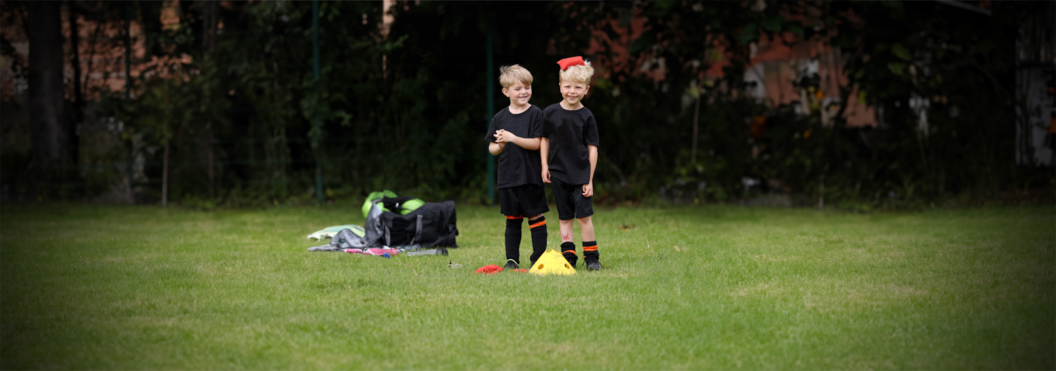 Two young boys stand next to a cone. One has a beanbag balanced on top of his head.