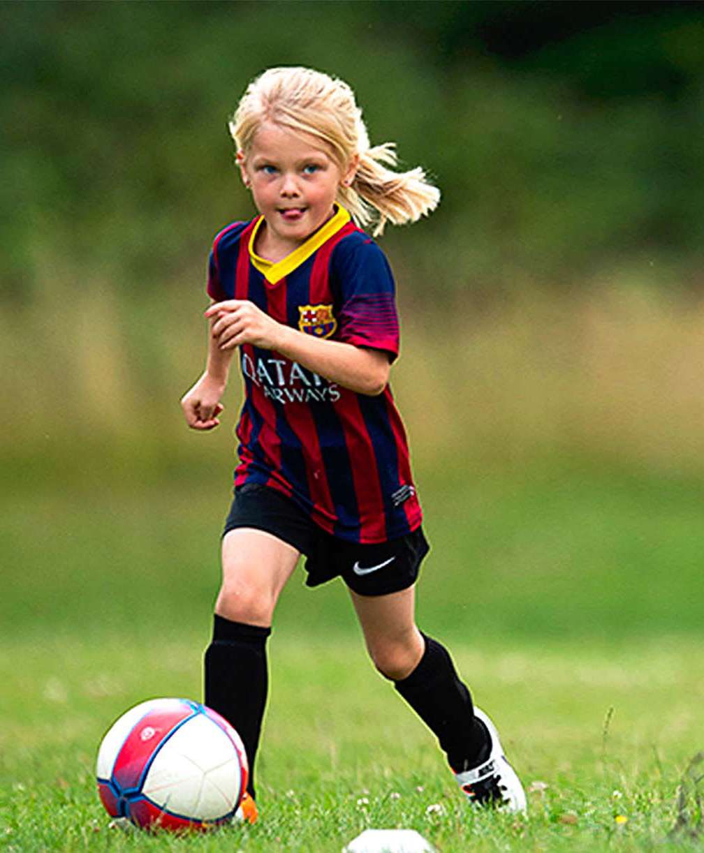 young girl with blonde hair kicks ball, she wears a FC Barcelona kit