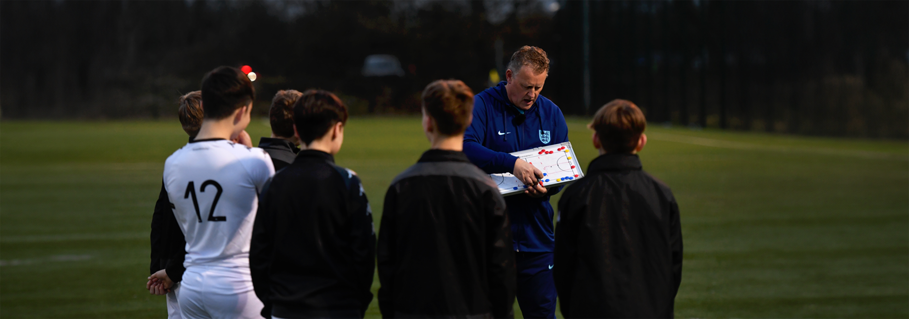 A small group of players gather around a coach who's holding a tactics board.
