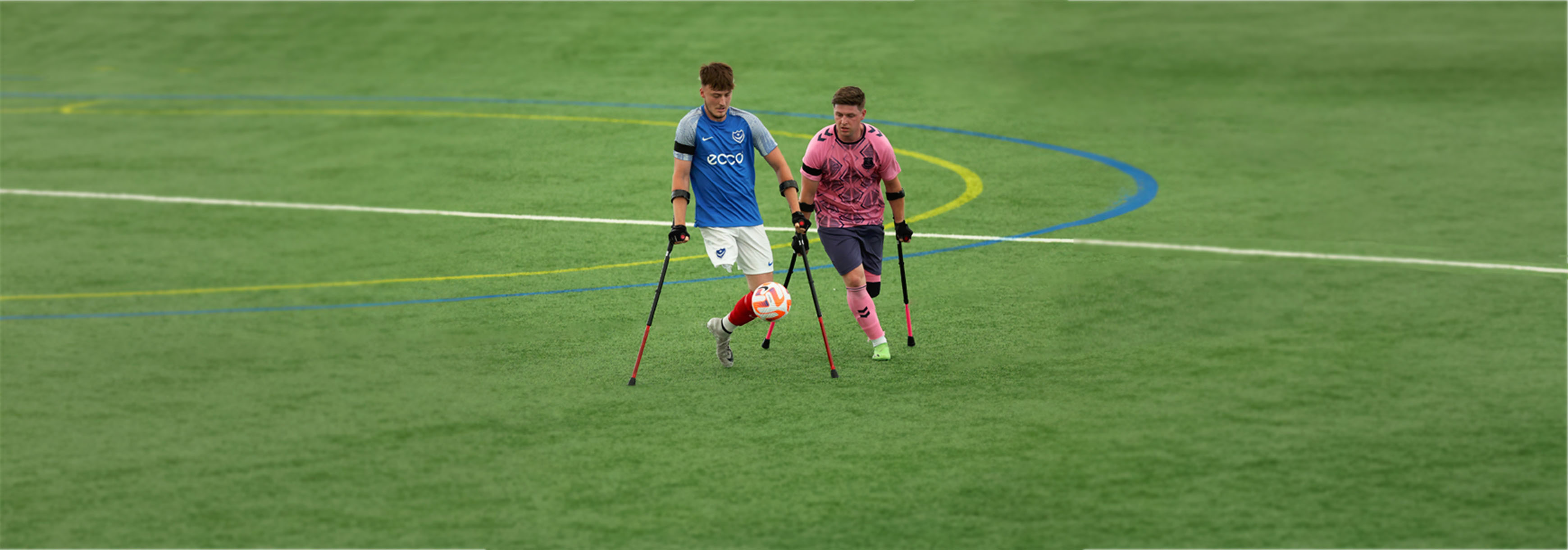 Two amputee players with crutches play football on an indoor 3G pitch.