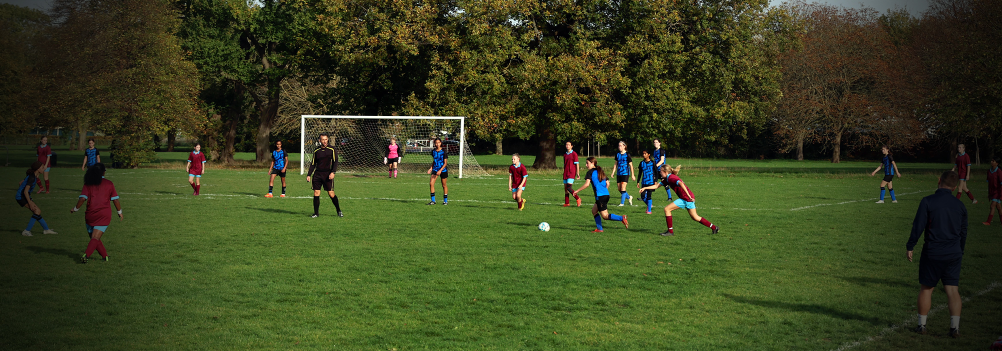 A wide-angle shot of a girls' grassroots football match. A player in a blue kit cuts inside with the ball with an opponent close behind her.