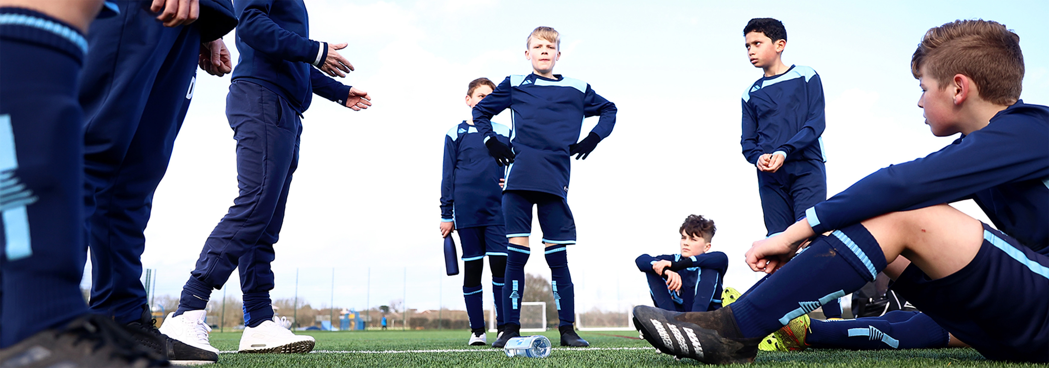 A group of boys listen to their coach at the side of a pitch on matchday. Some are standing while others are sat down.
