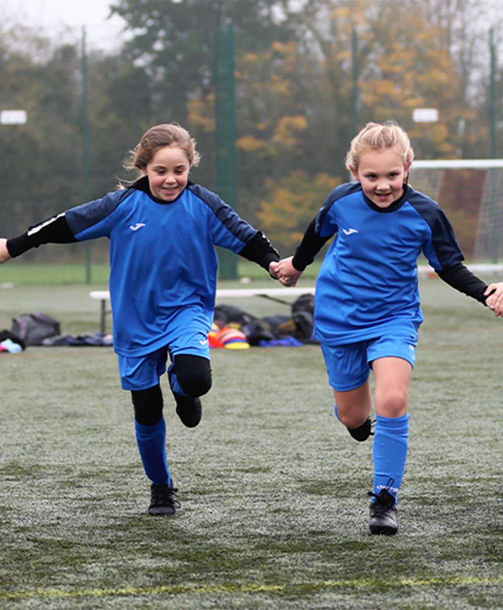 A group of young children run towards camera holding hands, dressed in football kit