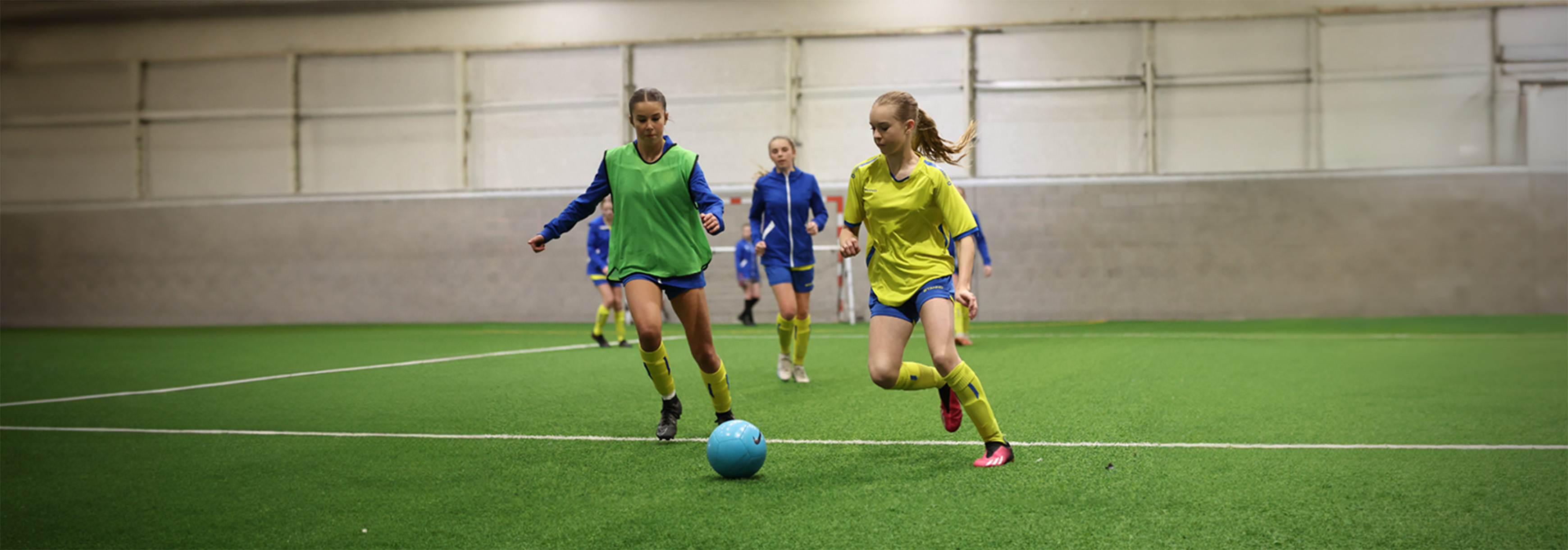 A girl in a yellow and blue kit runs with the ball under pressure from a green bibbed opponent to her right.