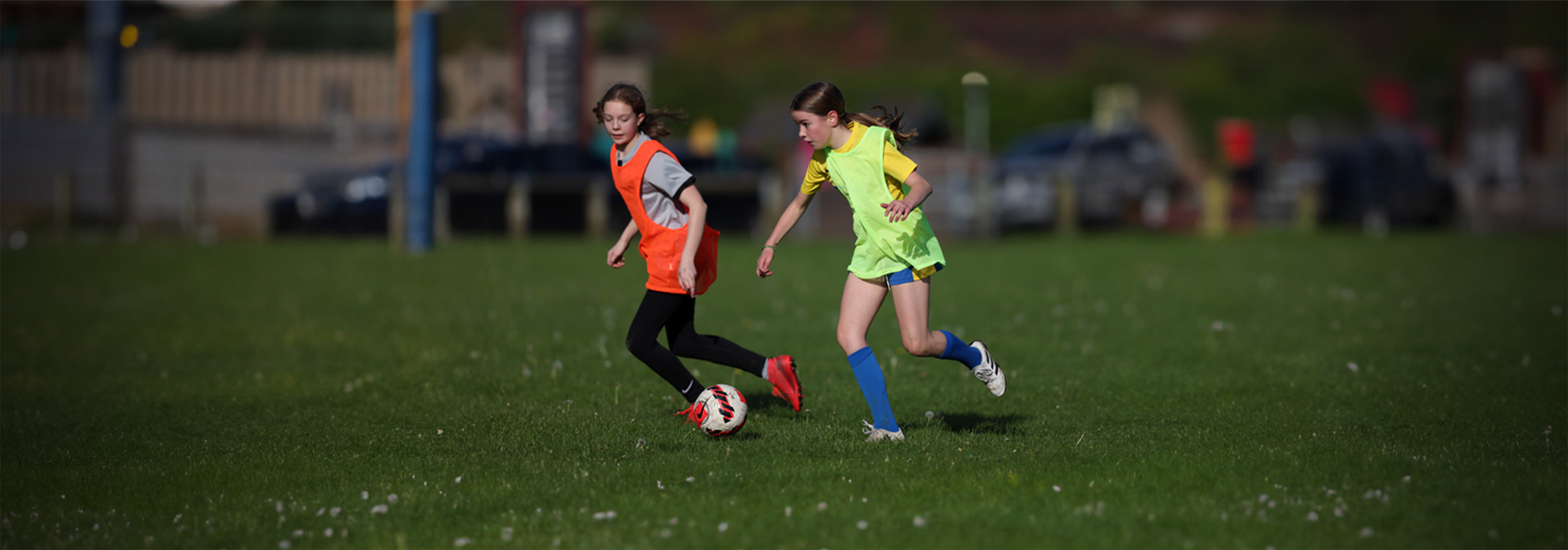 A young girl in a yellow bib runs forward with the ball on a grass pitch, with an orange-bibbed opponent to her right.