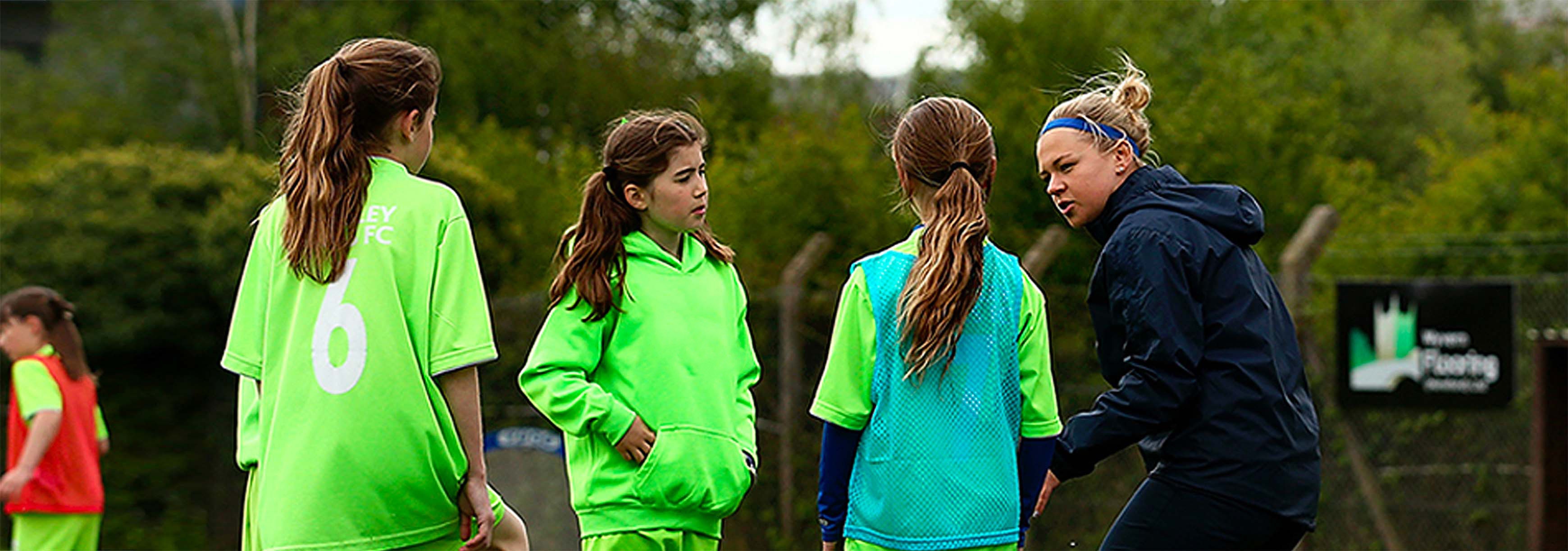 3 young female players wearing green kits look at their coach, a female wearing black. 