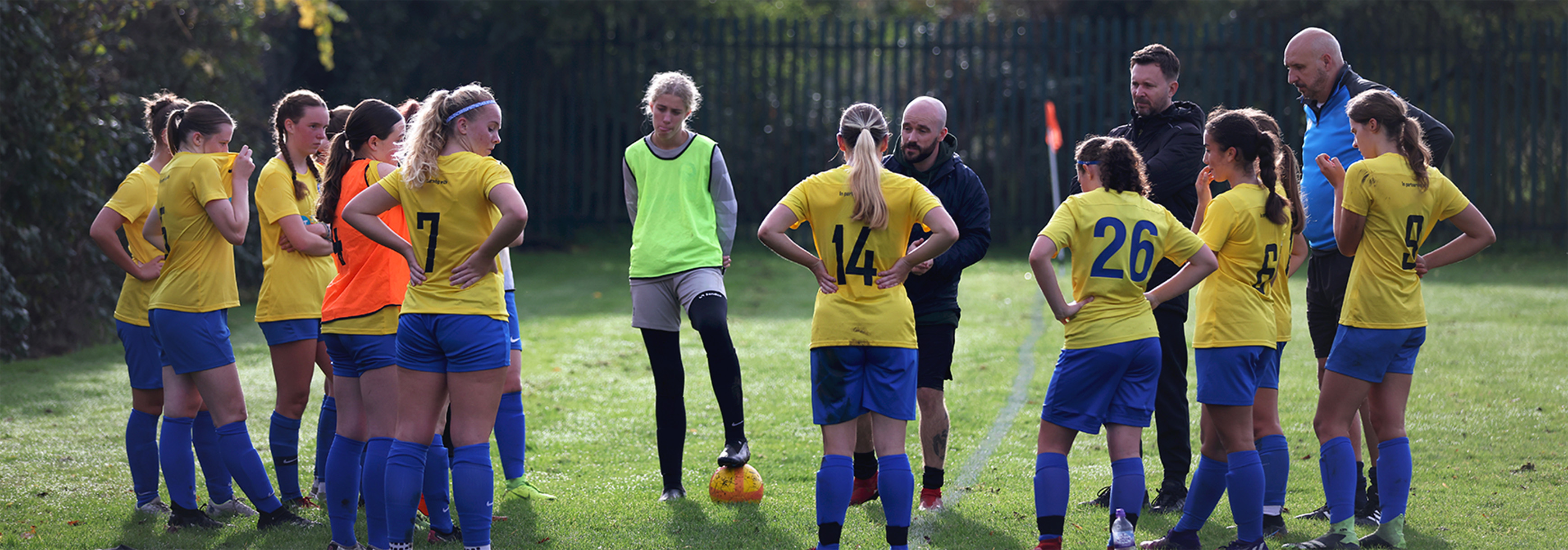 A team wearing yellow tops and blue shorts and socks stand in a circle to listen to their coach.