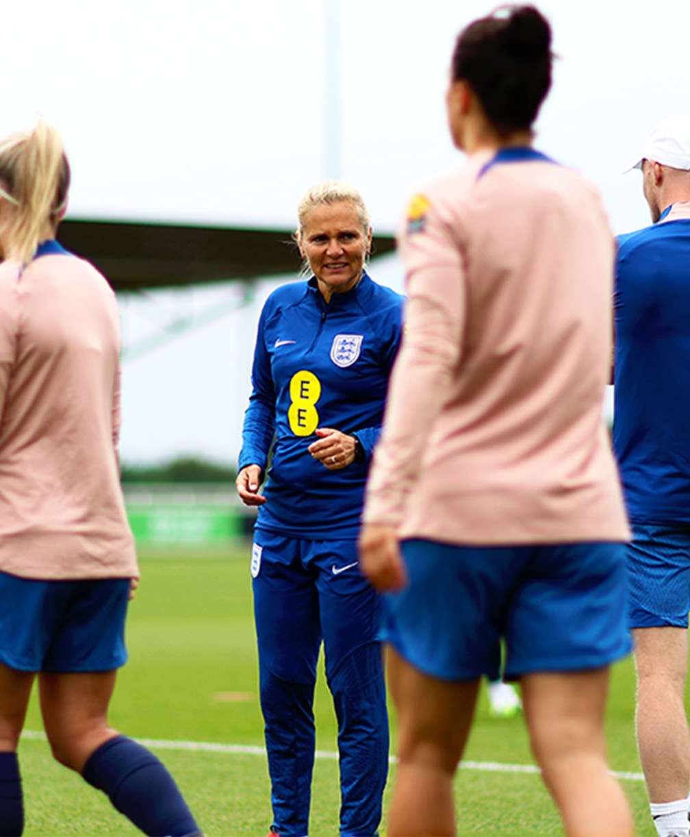 During training as players are walking onto the pitch at St. George's Park, England women’s head coach, Sarina Wiegman, slightly leans forward to listen to her assistant coach.