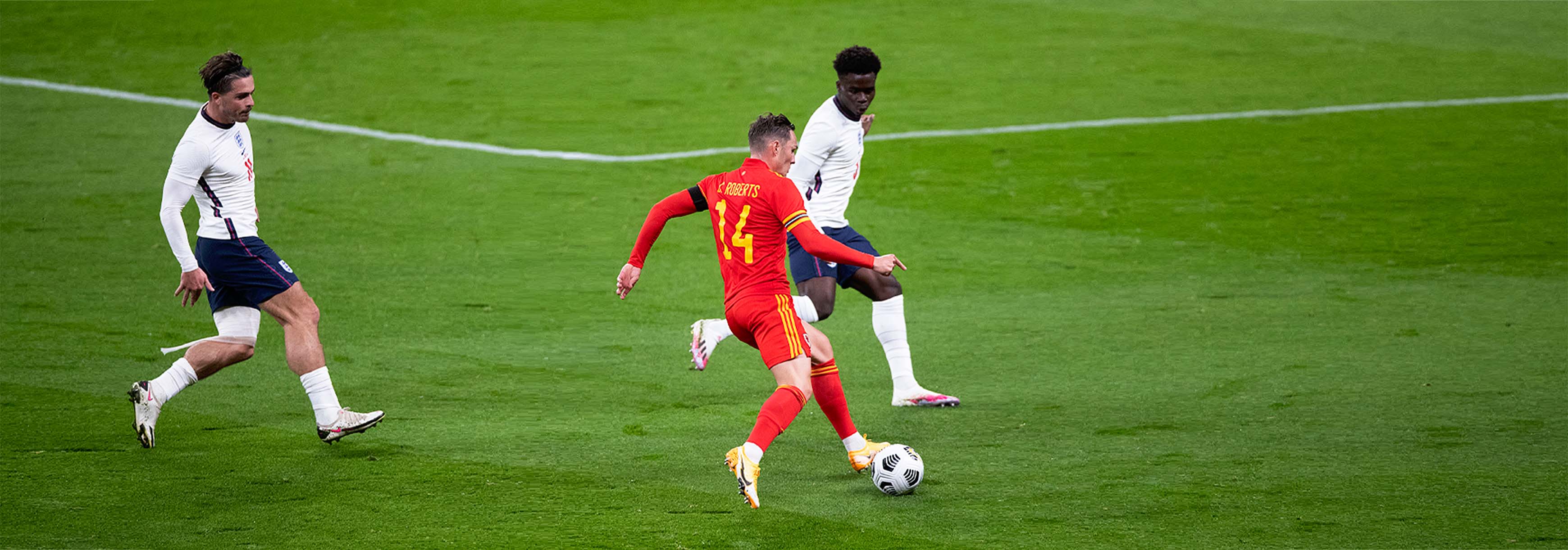 2 england players wearing white playing against a player from Wales wearing red. The player in red is going to kick the ball with his back faced to the camera.