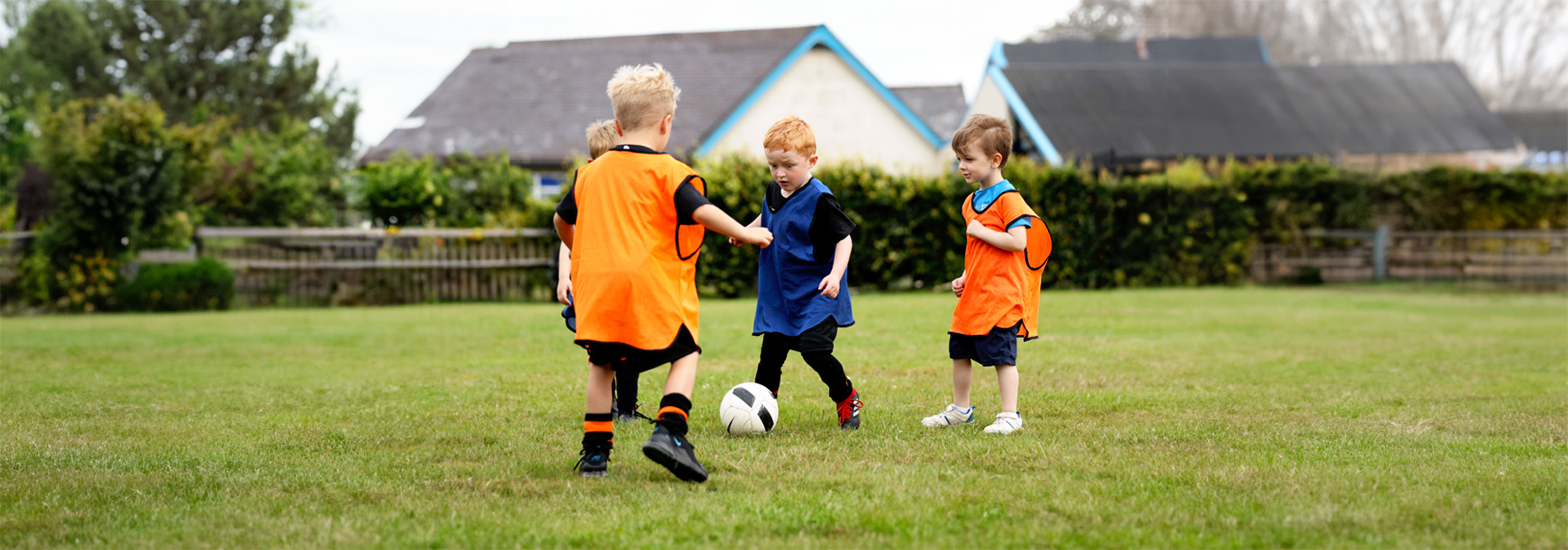 A group of four young children play football. One is kicking the ball, wearing a blue bib, while a teammate and two opponents stand round him.