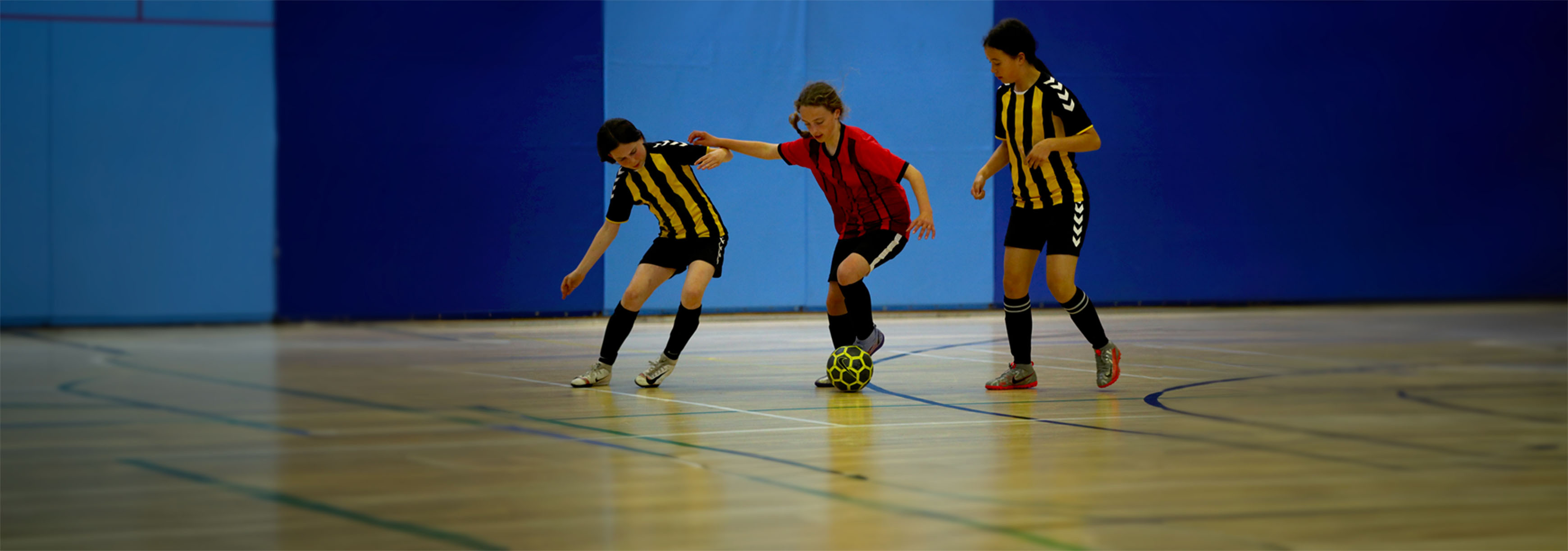 A girl runs with the ball while under pressure from two opponents during a futsal match in an indoor sports hall.