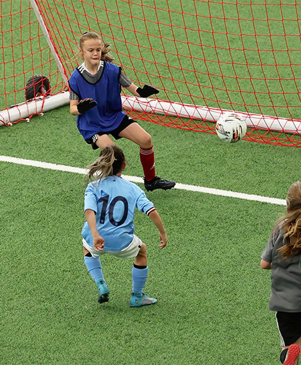 A young player uses the inside of her foot to send an effort towards goal from close range during a match on an indoor 3G pitch.