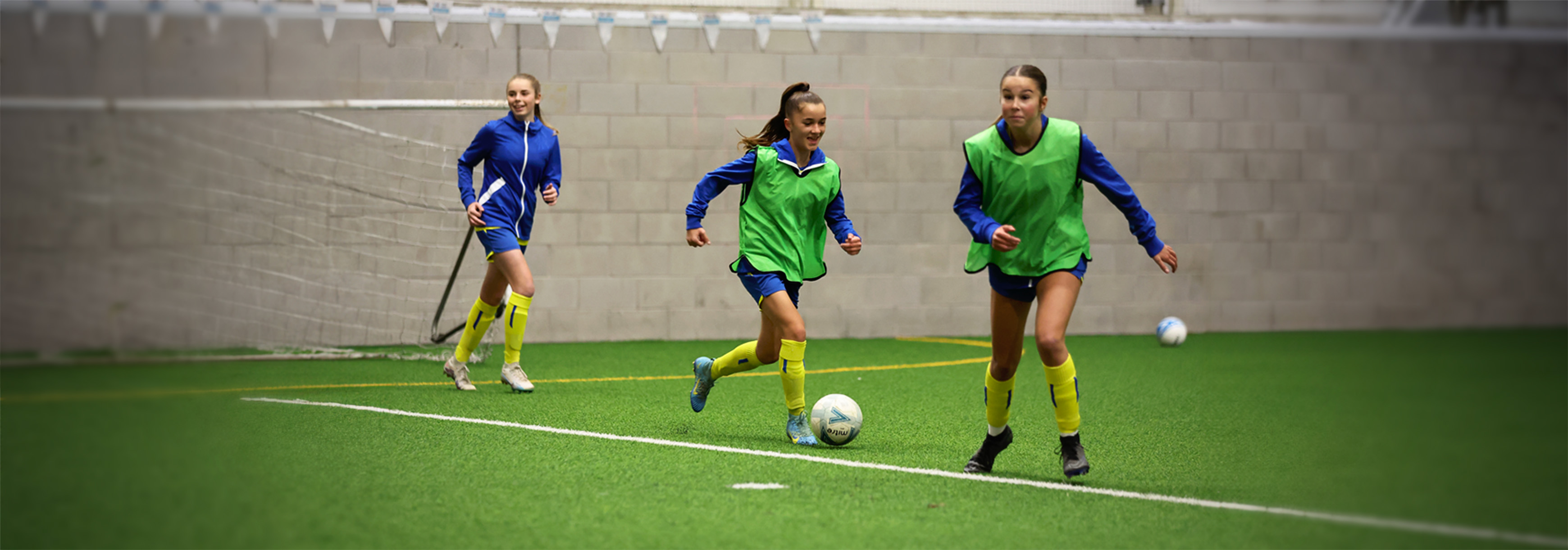 During training on an indoor pitch, one girl runs forward with the ball while two others move forward looking to receive a pass.