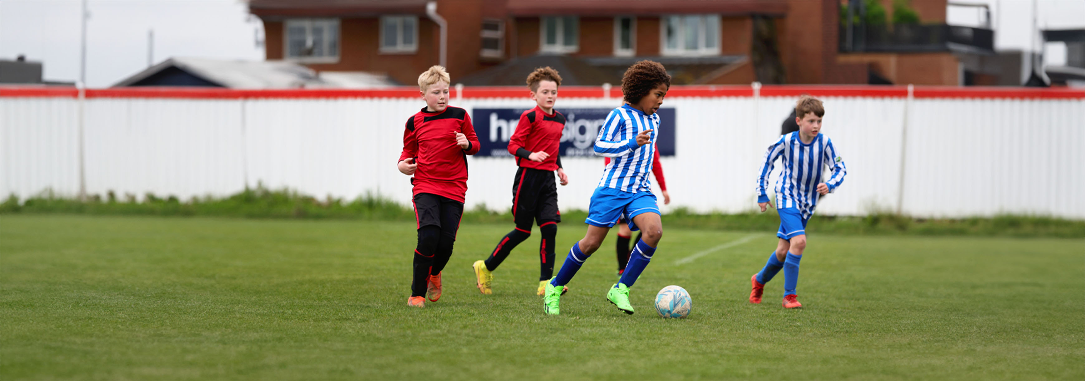 Young player wearing a blue and white striped kit runs with the ball. Beside him runs another child in the same kit, and behind are two players running wearing red and black kit.
