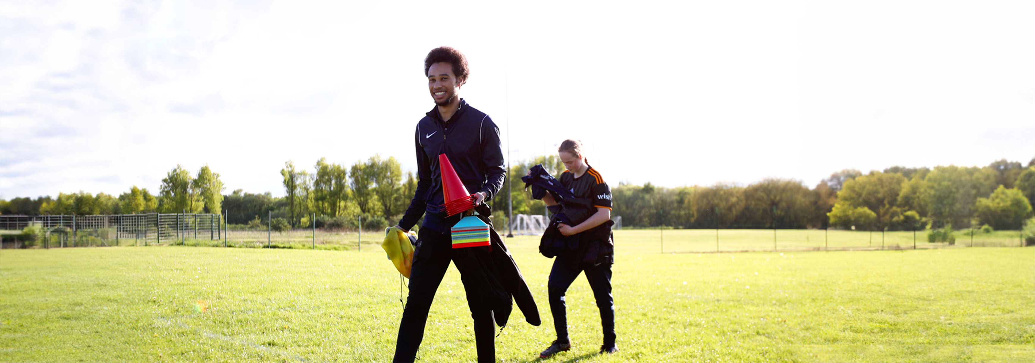 A coach walks onto a grass pitch holding some cones and a bag of bibs.