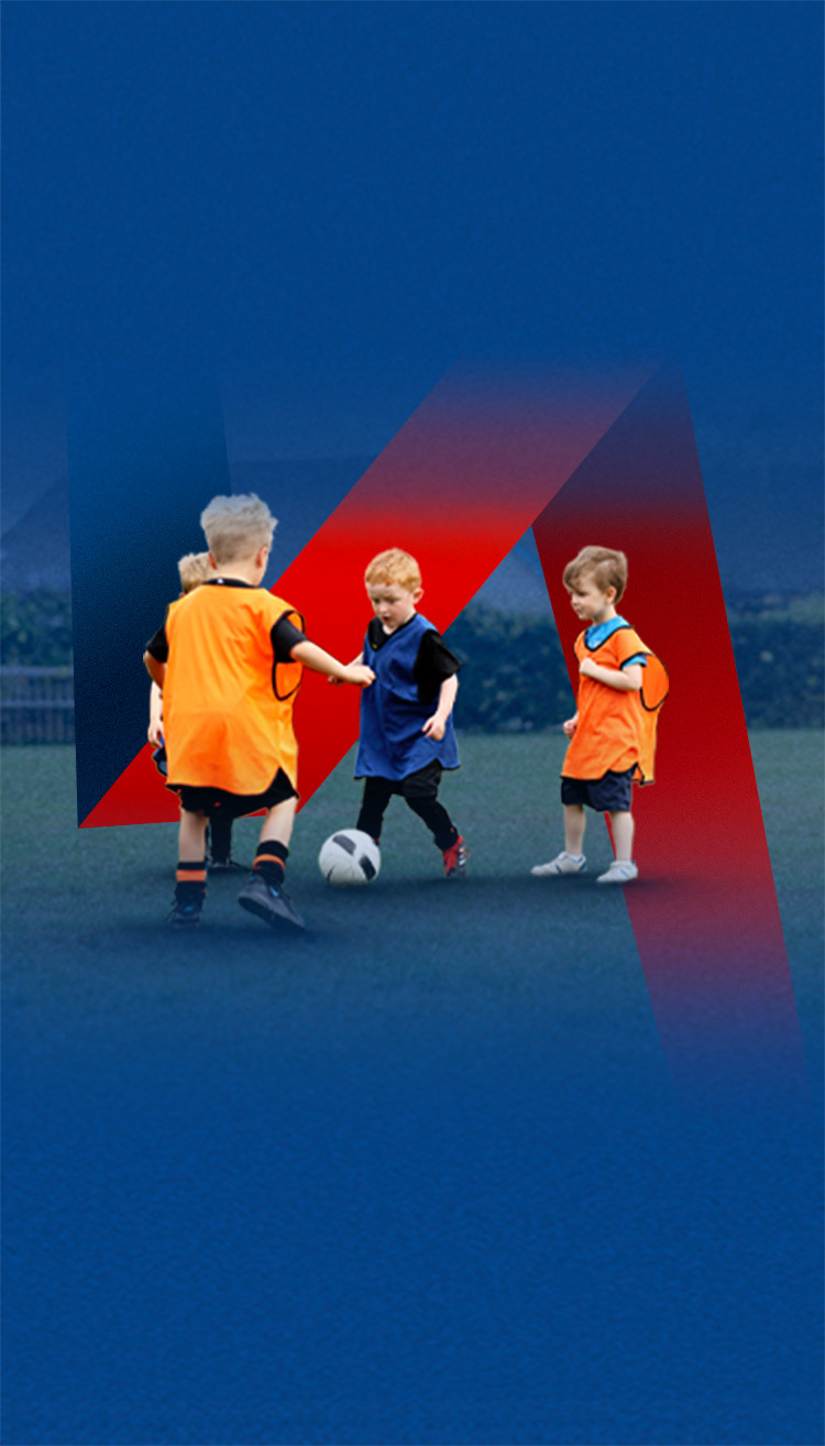 A group of four young children play football. One is kicking the ball, wearing a blue bib, while a teammate and two opponents stand round him.