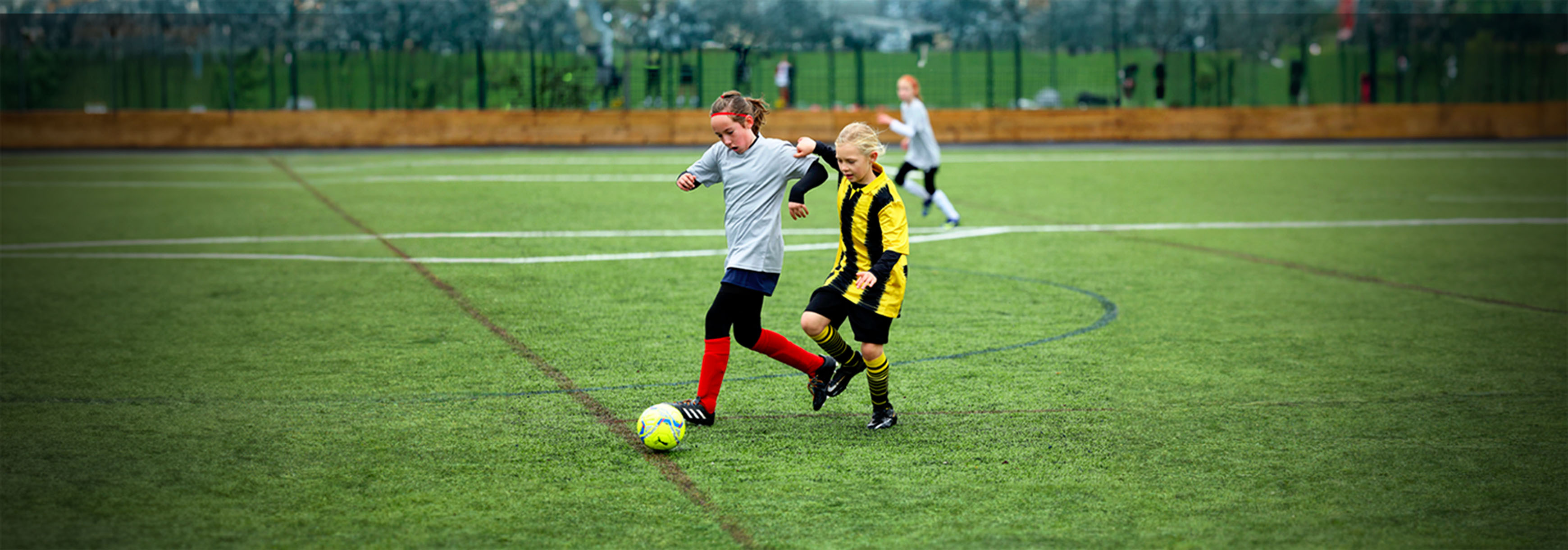 A girl runs with the ball while under pressure from an opponent on her left, during a match on a 3G pitch.