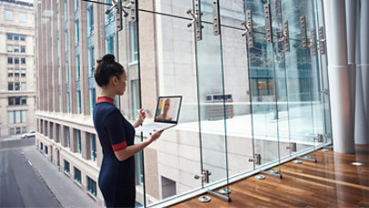 A businesswoman stands in front of a window holding a laptop and conducting a Teams call.