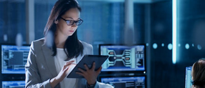 A woman with glasses reviews data on a tablet in a dimly lit tech control room.