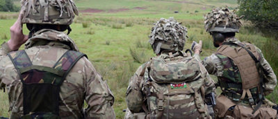 Three soldiers in camouflage gear standing in a field, facing away from the camera, observing the landscape.