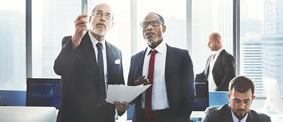 Two businessmen discussing over documents in a high-rise office, another man working in the background.