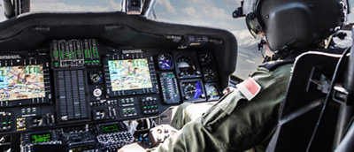 Pilot in military helicopter cockpit with detailed instrument panels and multiple display screens.