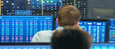 Back view of two people monitoring financial data on multiple blue screens in a control room environment.