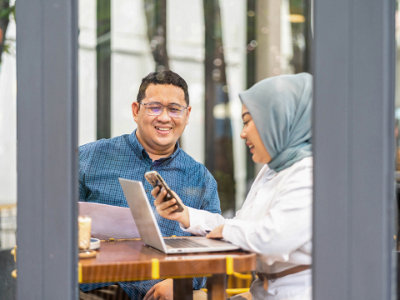 Two people sit in a cafe, smiling and working on a project. One person in a blue shirt holds a document, while the other in a hijab.