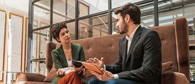 Dois profissionais discutindo em um tablet digital enquanto estão sentados em um sofá em um lobby de escritório moderno.