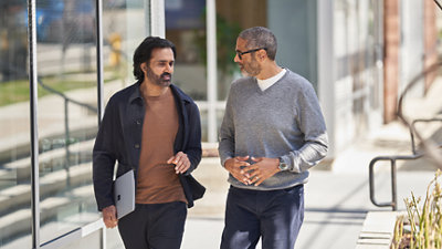 Two men walking outside, engaged in conversation. One is holding a laptop, and they are passing by glass windows.