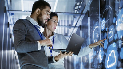 Two professionals analyzing data on screens in a high-tech server room, one holding a laptop