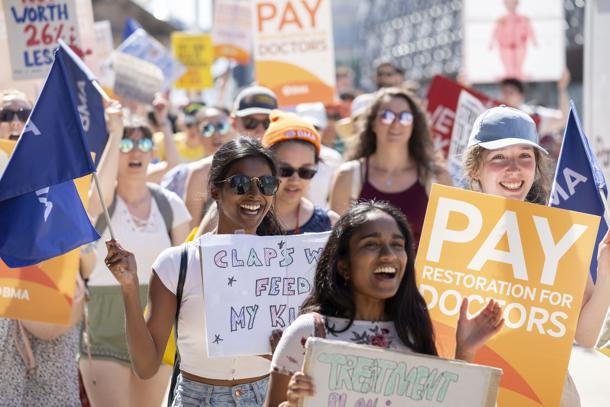 A group of smiling doctors march, hold handmade and BMA placards and wave flags in the sun outside at a junior doctor pay restoration march that took place on 15 June 2023