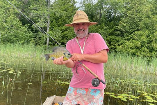 A smiling man wearing a straw hat holds a fishing rod and a bluegill sunfish.