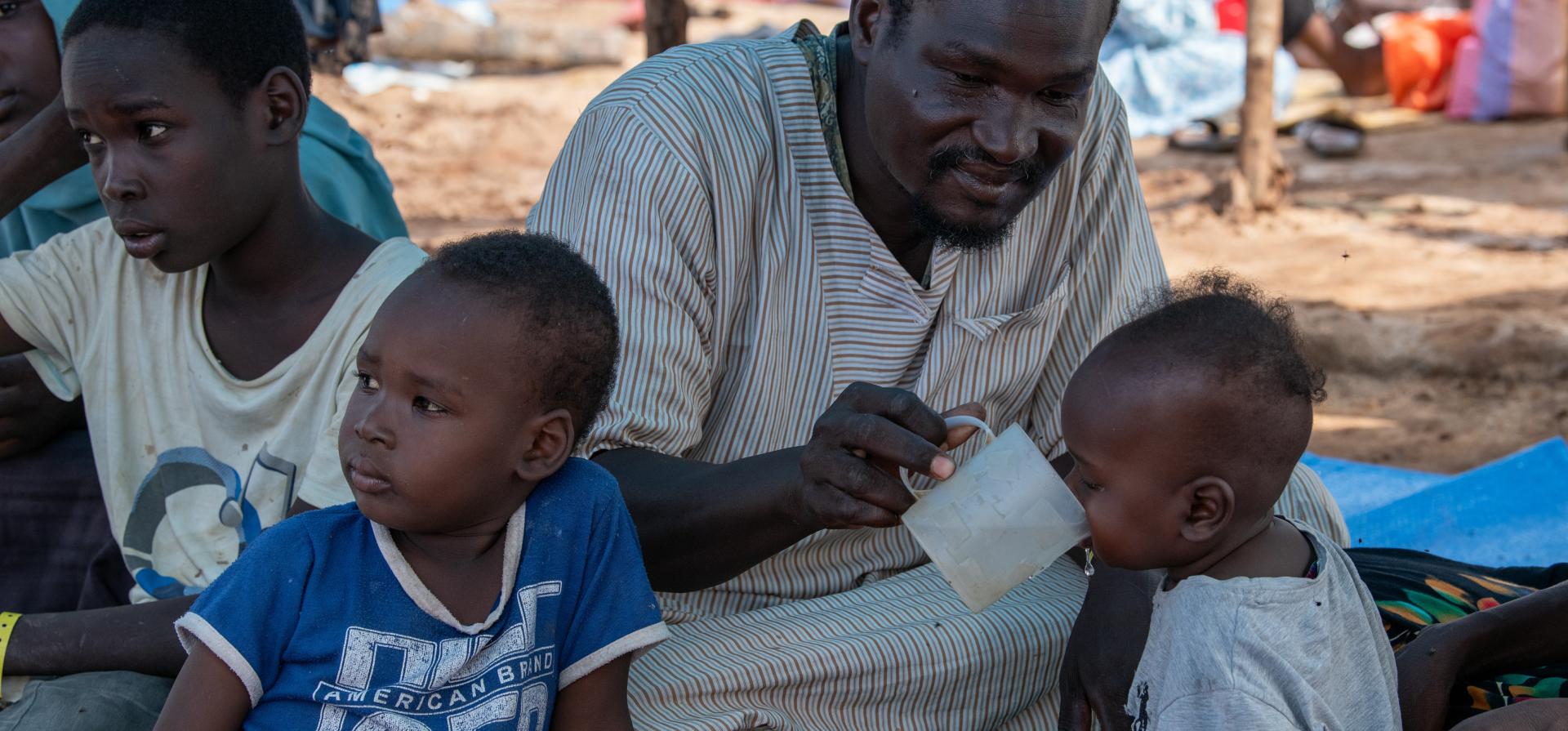 A group of four people, three children and one man sit in the shade. The man helps the youngest child to drink from a cup.