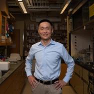 Prof. Chuan He faces camera smiling with hands on hips with a chemistry lab in the background